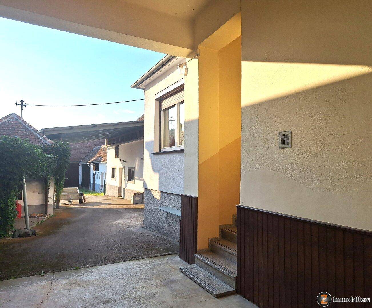 Eingangsbereich eines Gebäudes mit Treppe unter einem schützenden Vordach im Sonnenlicht.