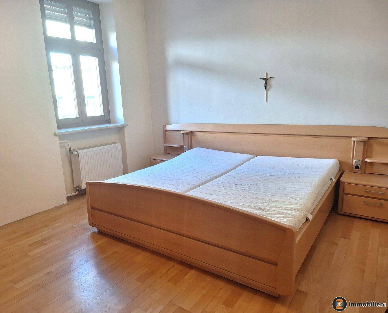 Schlafzimmer mit hellem Holzbett, Parkettboden und einem Fenster mit Heizkörper darunter.