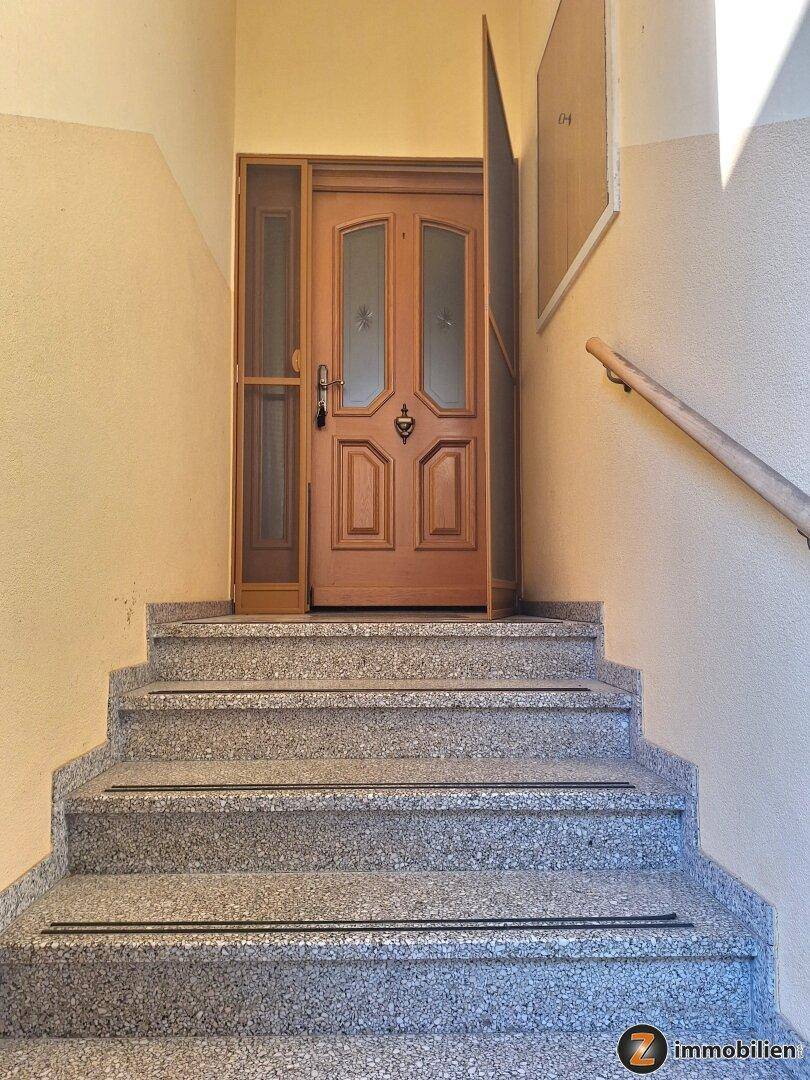 Heller Flur mit Steintreppe, die zu einer hölzernen Eingangstür führt.