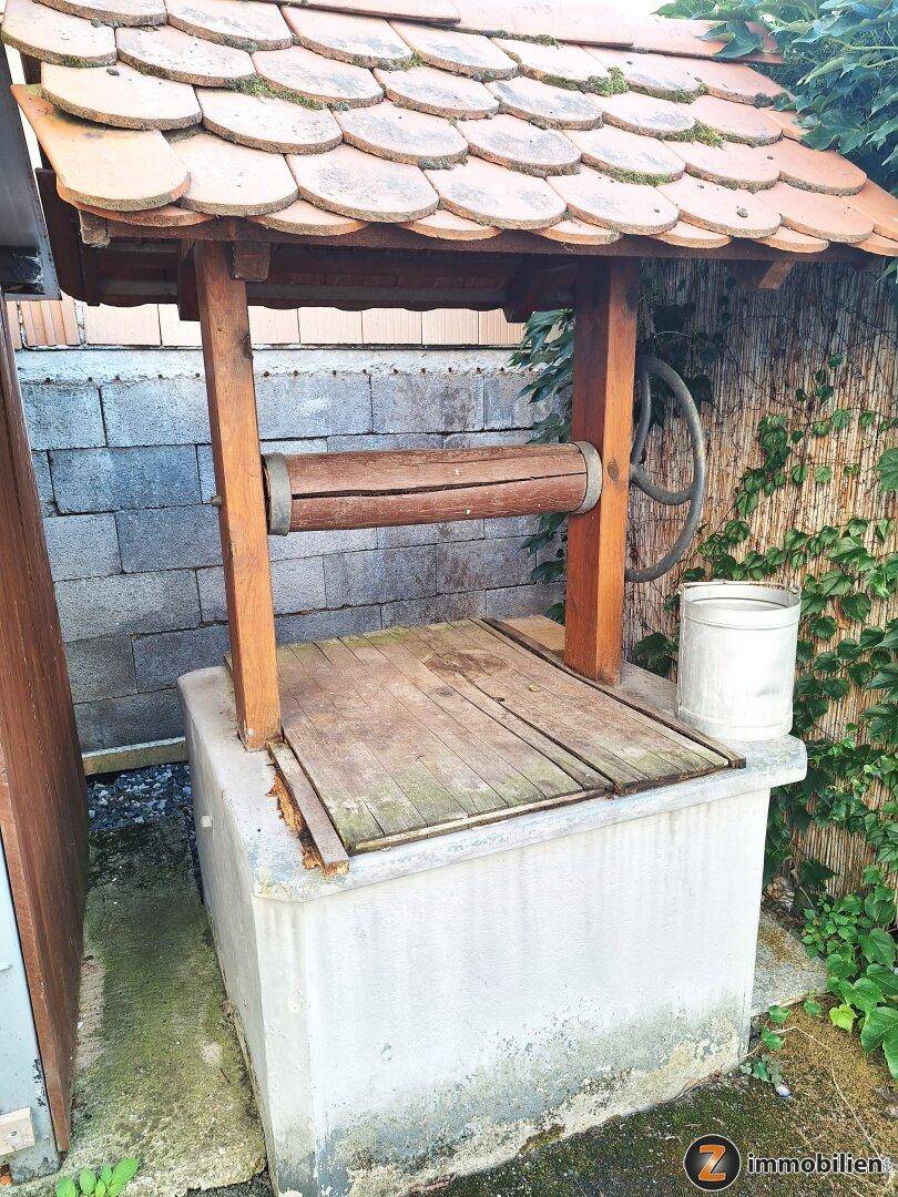 Nahaufnahme eines traditionellen Brunnens mit Holzdach und gemauertem Sockel im Außenbereich.