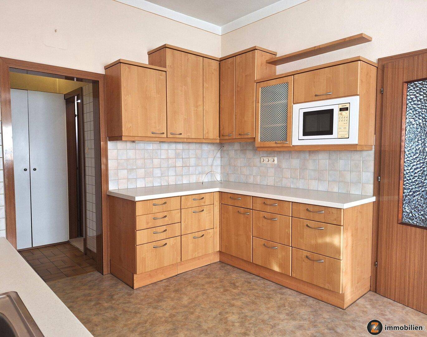 Küche mit hellen Holzschränken, weißen Arbeitsplatten, Fliesenrückwand und integrierter Mikrowelle.