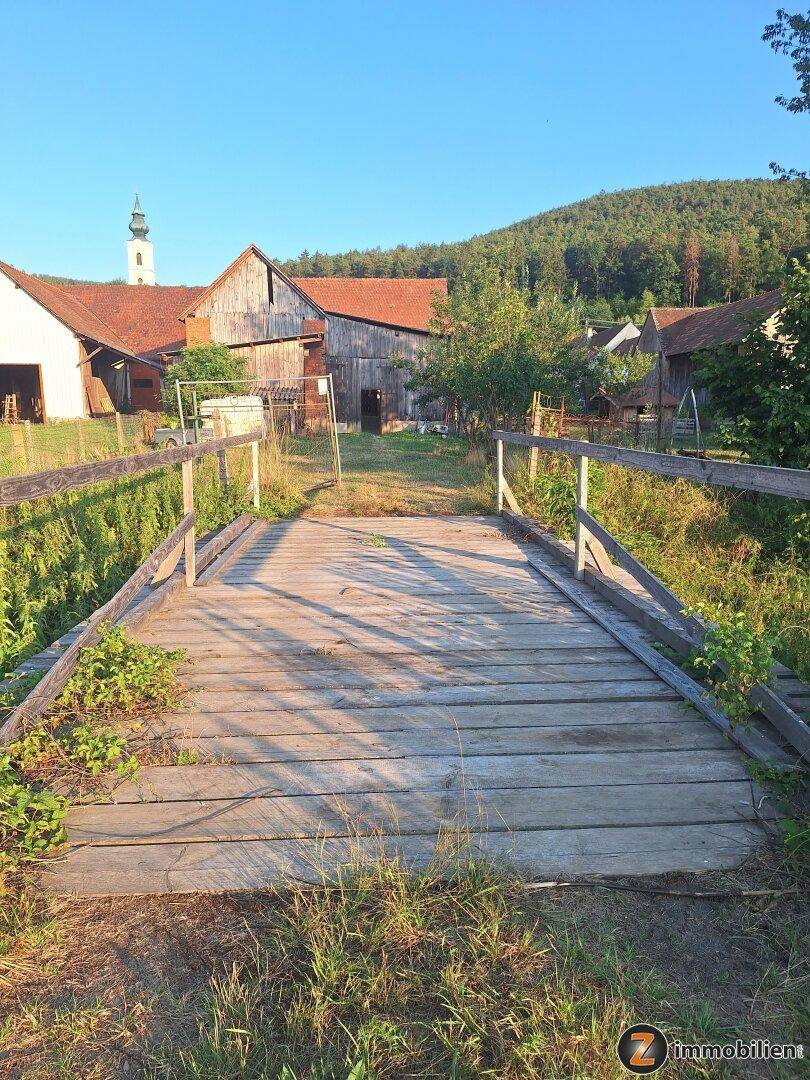 Holzbrücke führt zu einem ländlichen Anwesen mit einem Kirchturm im Hintergrund.