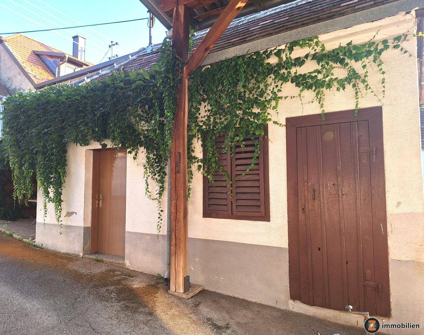 Fassade eines kleinen Gebäudes, teilweise mit Efeu bewachsen, mit Holztür und Fensterläden.