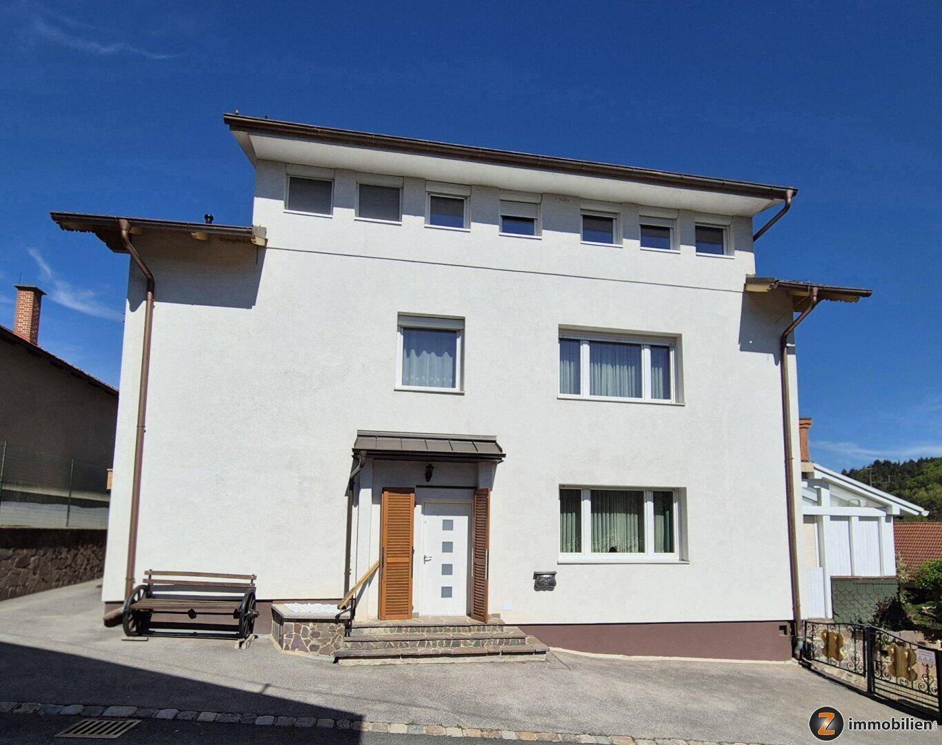 Vorderansicht eines Einfamilienhauses mit weißer Fassade, kleinen Fenstern und einem Vordach über dem Eingang.