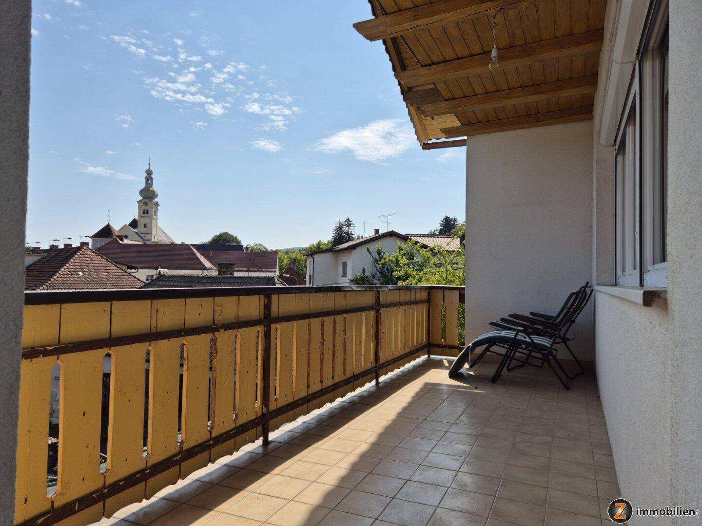 Sonniger Balkon mit Fliesenboden und weitem Blick über die Dächer und eine Kirche.