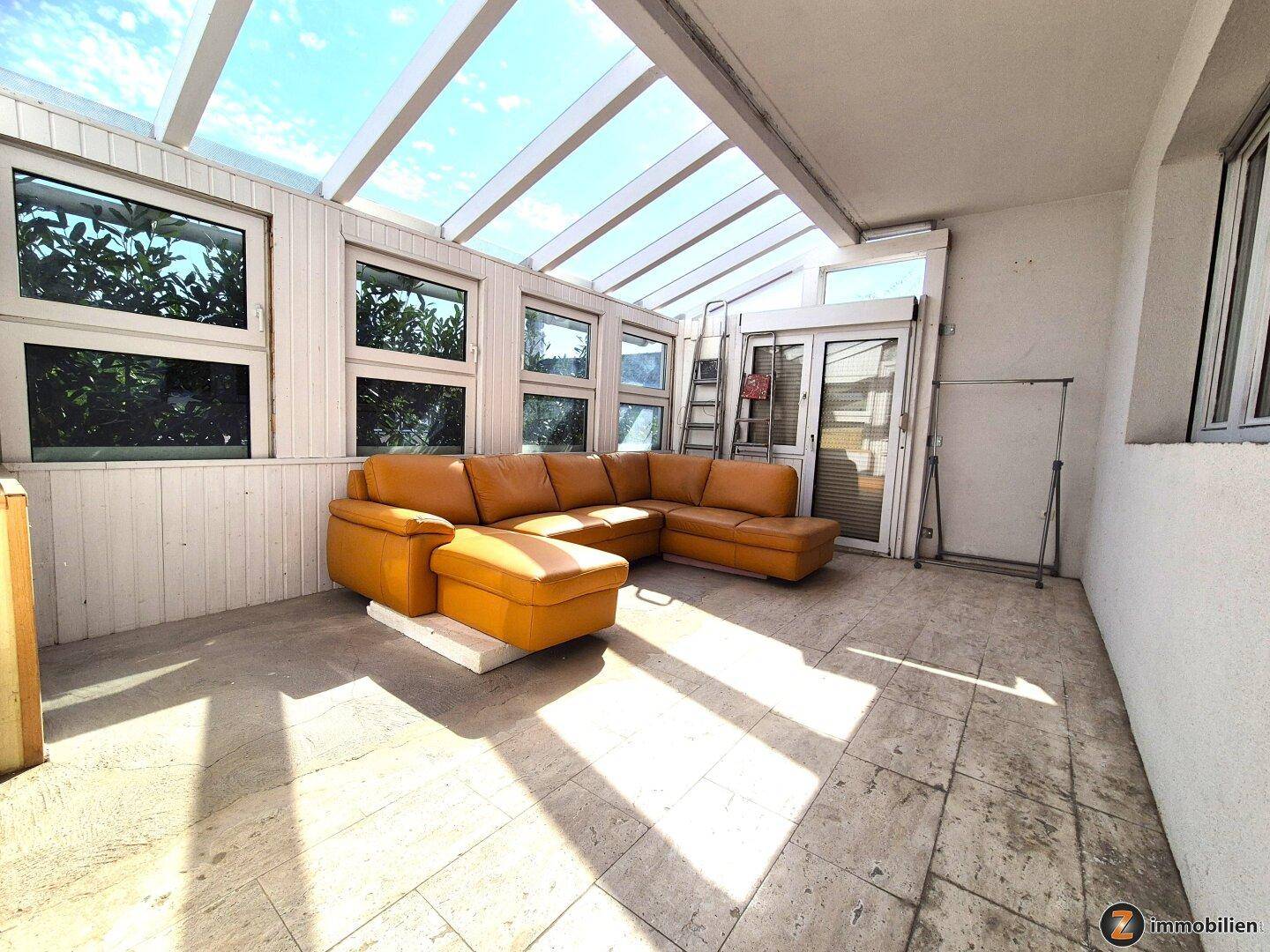Heller Wintergarten mit Fliesenboden, Ledersofa und viel Tageslicht durch das Glasdach.