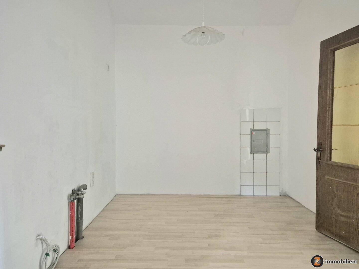 Leerer Raum mit Laminatboden, weißen Wänden und einem Kachelofen-Element.