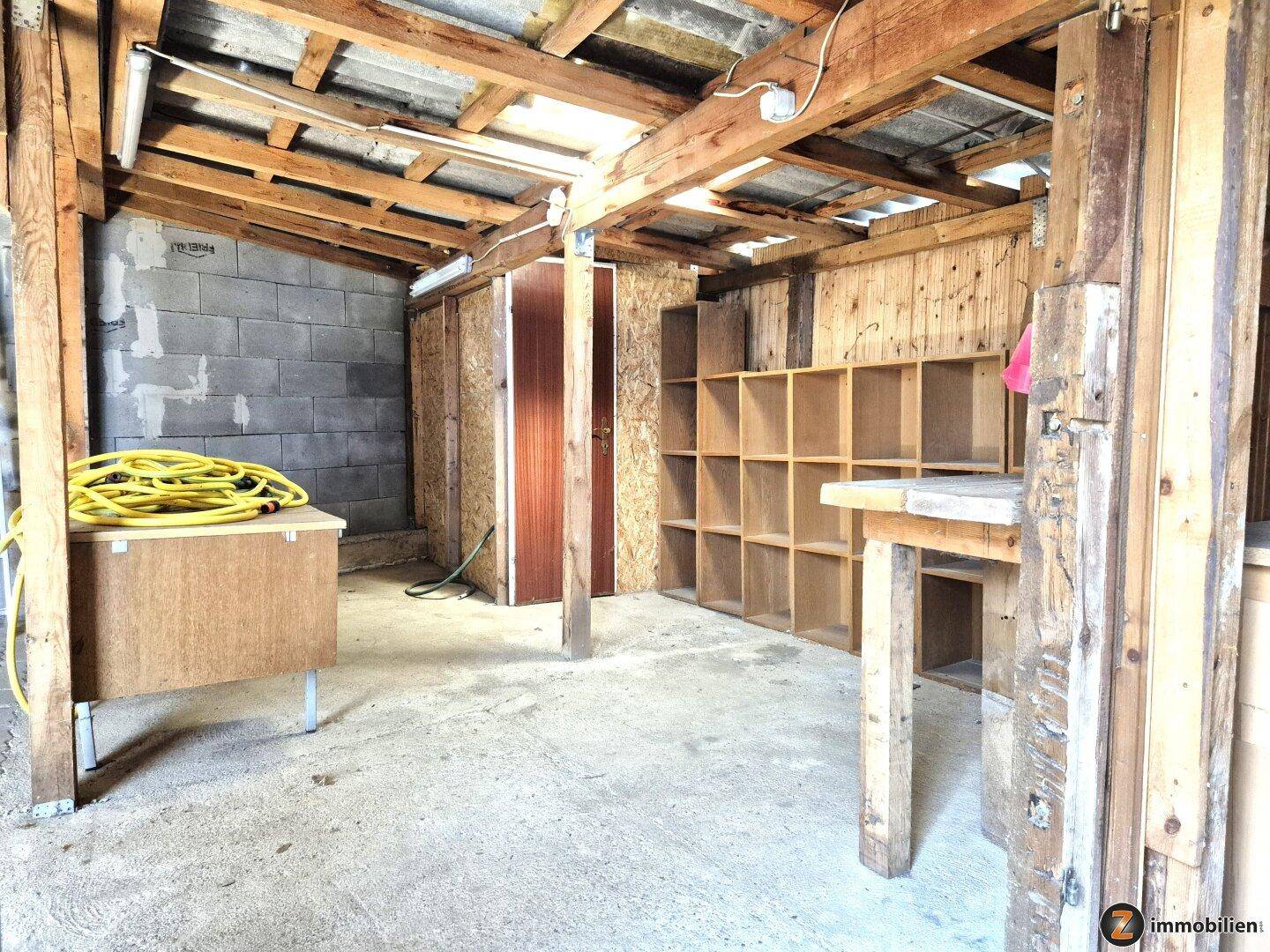 Einfacher Lagerraum mit Holzregalen, Betonwänden und sichtbaren Holzbalken der Dachkonstruktion.