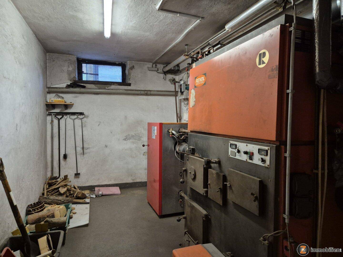 Heizungsraum mit großem Heizkessel, gestapelten Holzscheiten und einem kleinen Fenster zur Belüftung.