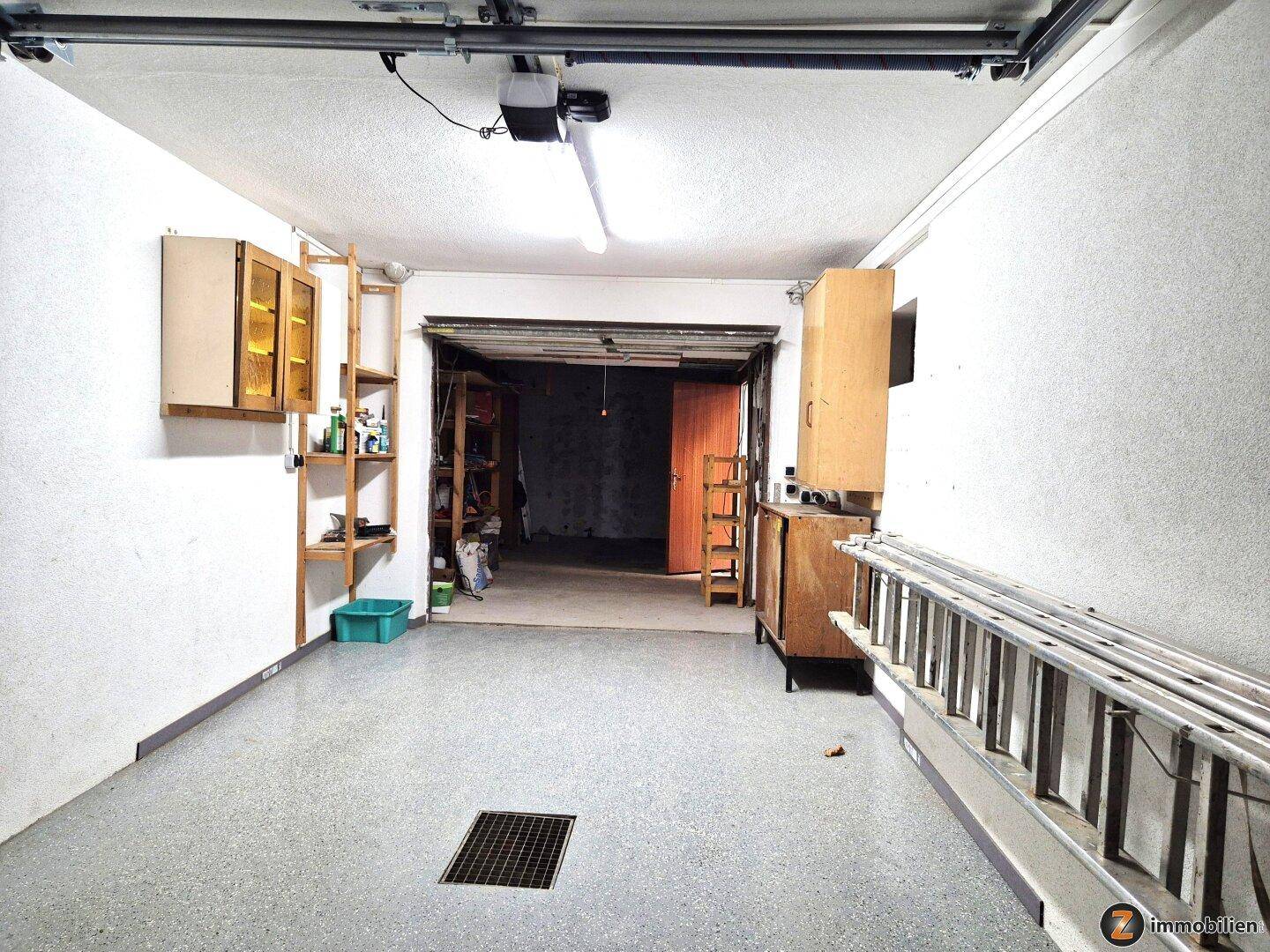 Geräumige Garage mit Betonboden, Regalen und offenem Tor zu einem weiteren Lagerbereich im Hintergrund.