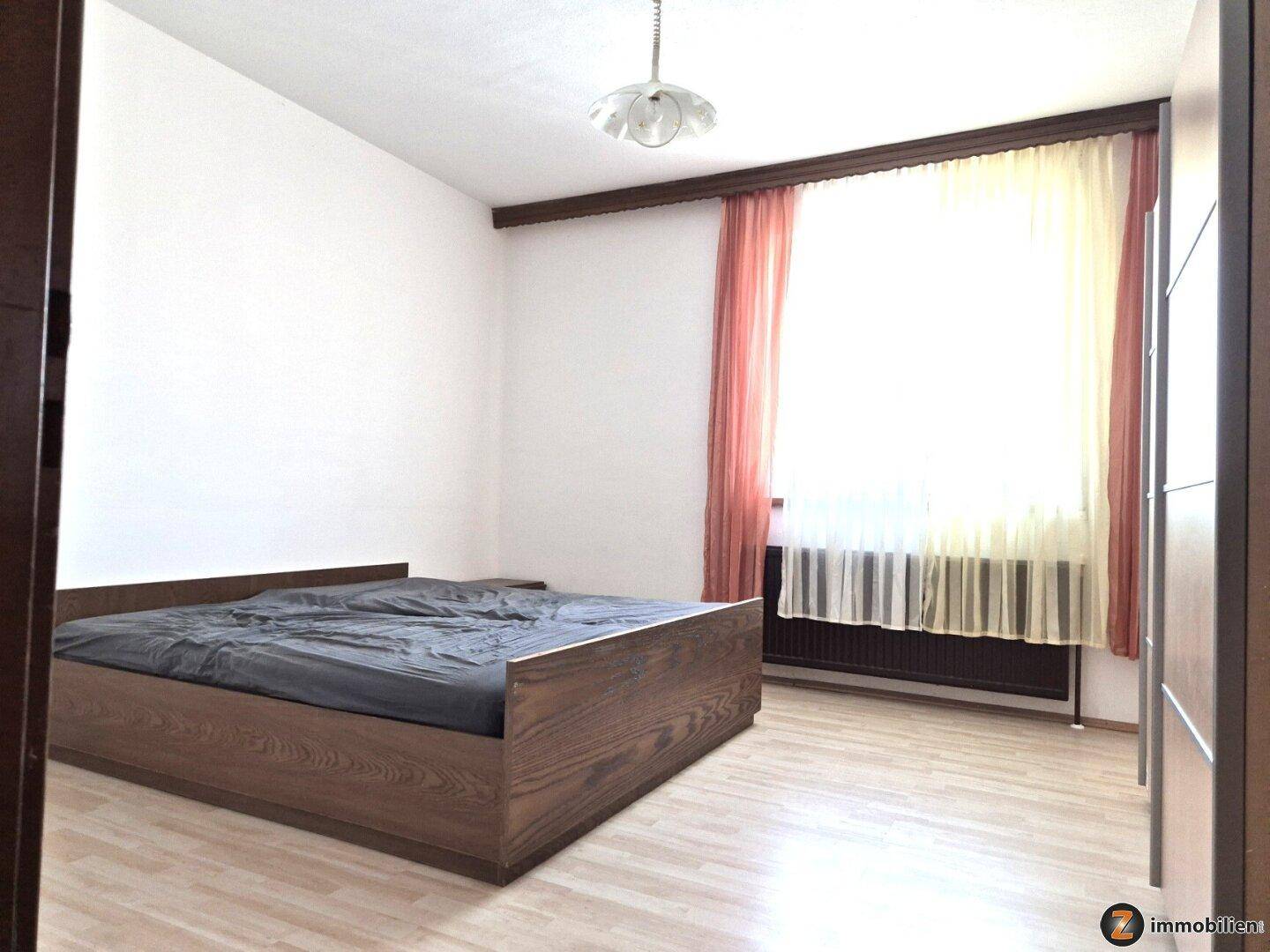 Schlafzimmer mit Doppelbett, Laminatboden und einem großen Fenster mit Vorhängen.
