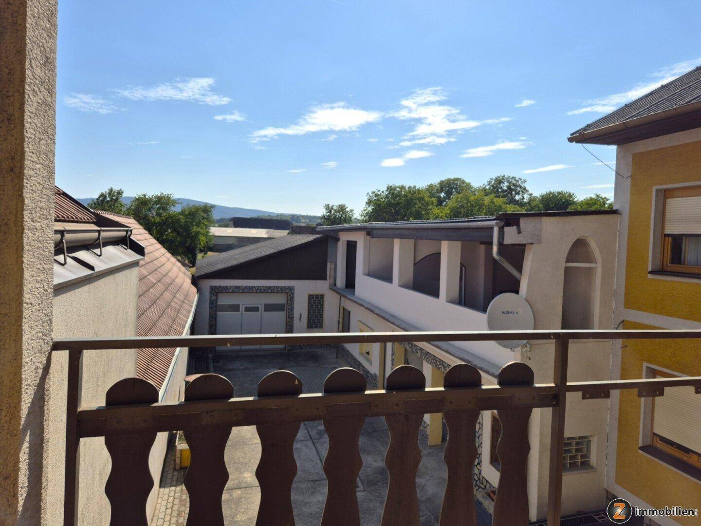 Blick vom Balkon auf umliegende Gebäude, eine Garage und die entfernte Landschaft unter blauem Himmel.