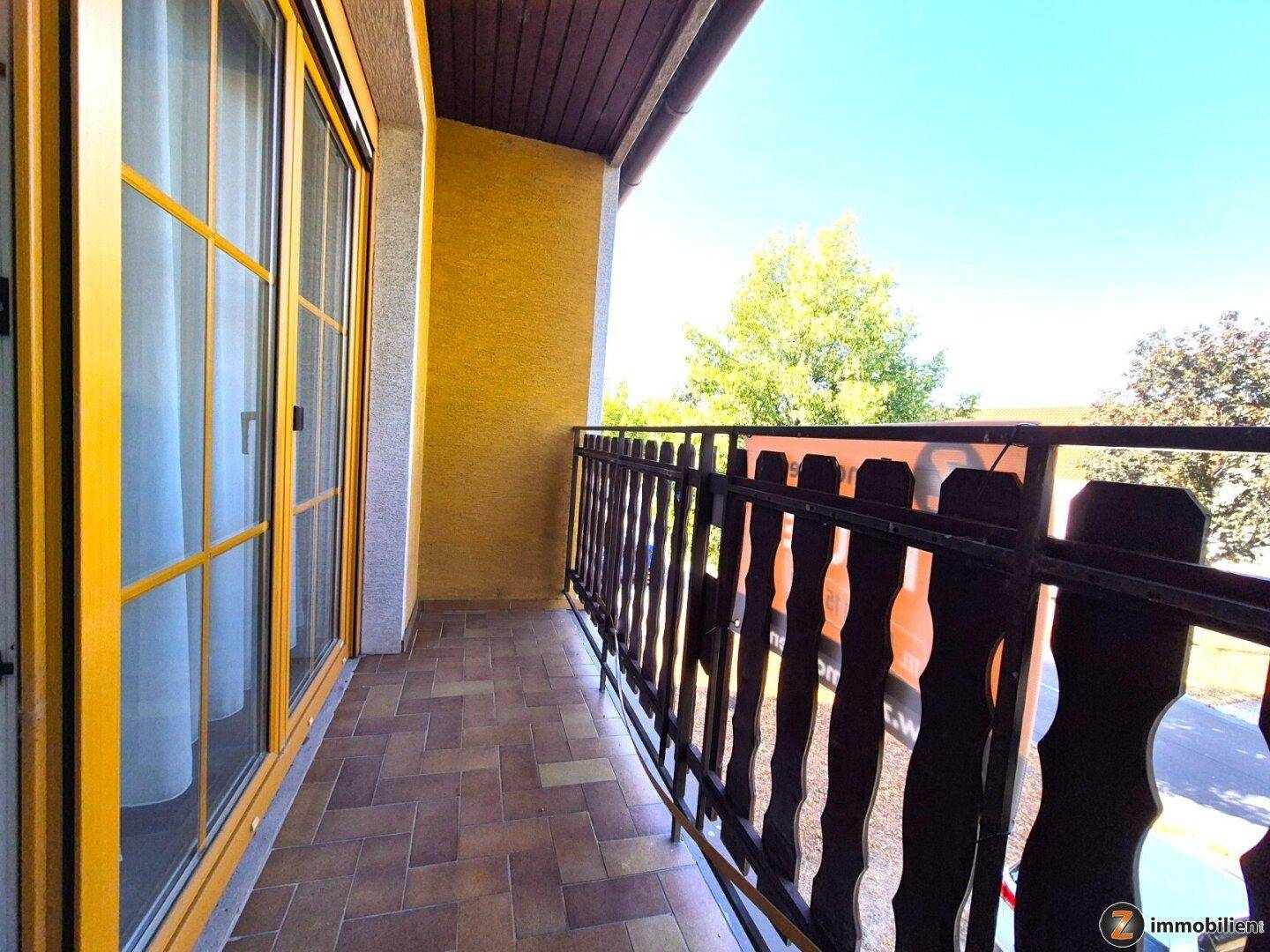 Heller Balkon mit braunen Fliesen, Holzgeländer und großer Glasschiebetür zum Innenbereich.