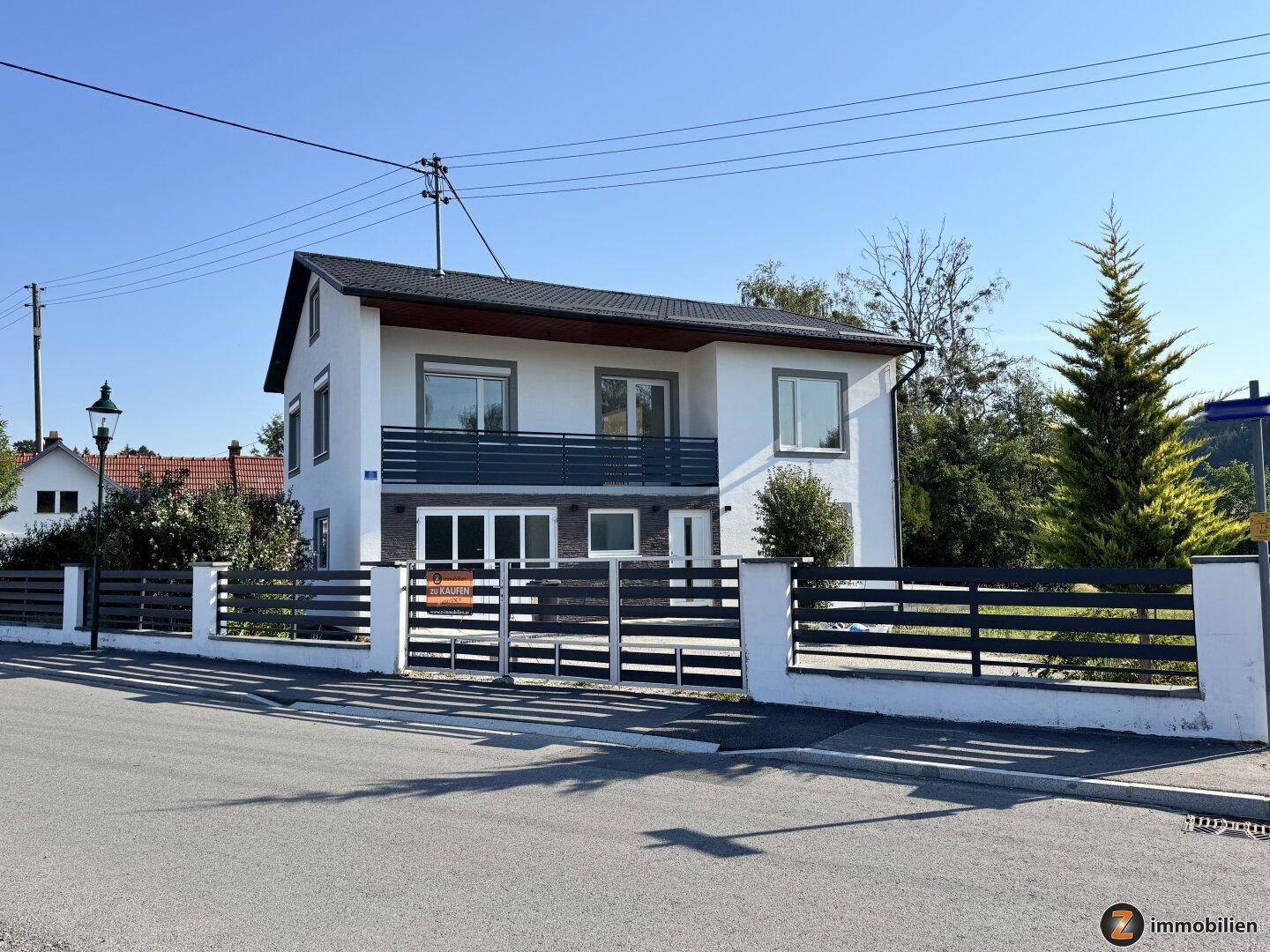 Freistehendes Einfamilienhaus mit weißer Fassade, Balkon und umzäuntem Vorgarten in sonniger Lage.