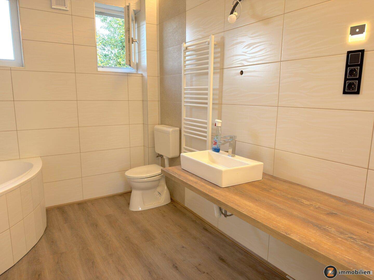 Modernes Badezimmer mit Eckbadewanne, Waschtisch und Handtuchheizkörper, hell gefliest.