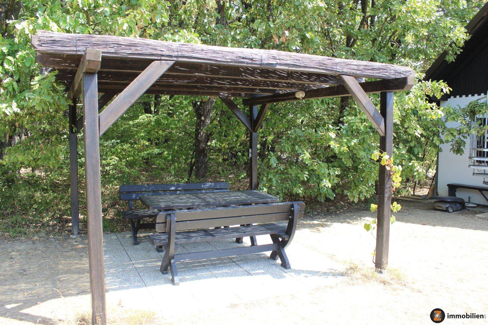Rustikale Holzpergola mit Sitzbänken im grünen Garten, umgeben von Bäumen.