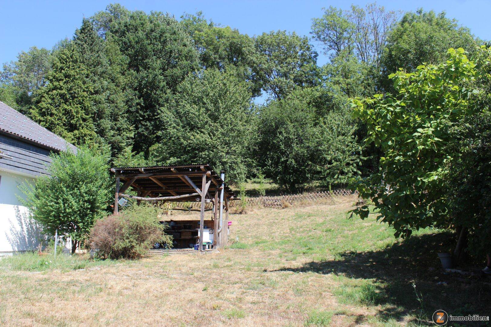 Großer Gartenbereich mit Holzunterstand und altem Baumbestand im Hintergrund.
