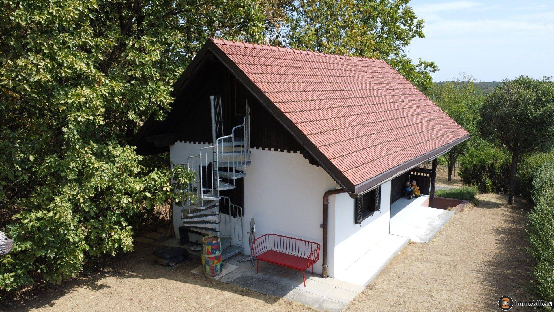 Seitenansicht des Hauses mit einer Außenspindeltreppe und rotem Ziegeldach, umgeben von Bäumen.