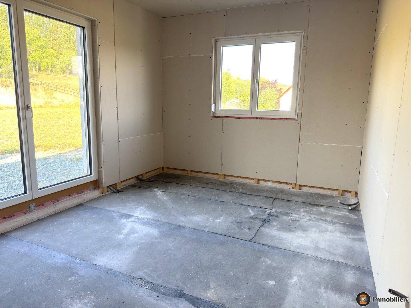 Rohbauzimmer mit Trockenbauwänden, Betonboden und zwei Fenstern für helle Atmosphäre.