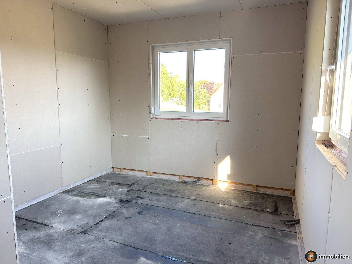Rohbauzimmer mit Trockenbauwänden, Betonboden und einem Fenster, das viel Licht hereinlässt.