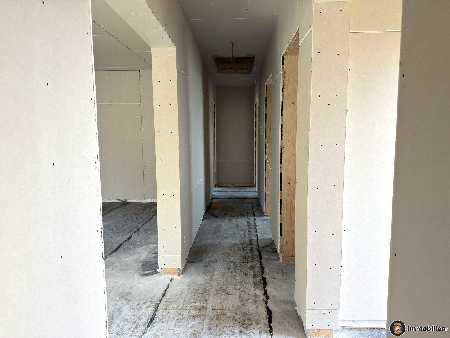 Flur im Rohbau mit Trockenbauwänden und Betonboden, der zu verschiedenen Räumen führt.