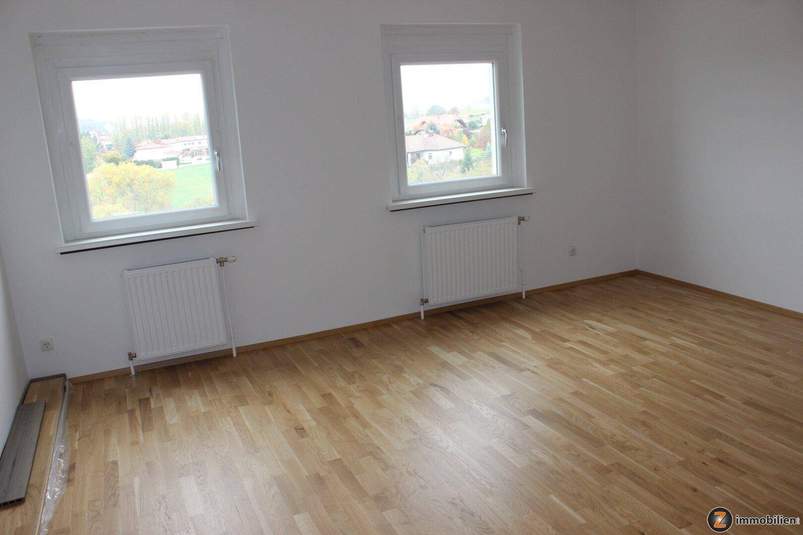 Leeres Schlafzimmer mit hellem Parkettboden, zwei Fenstern und Heizkörpern, bietet Blick ins Grüne.