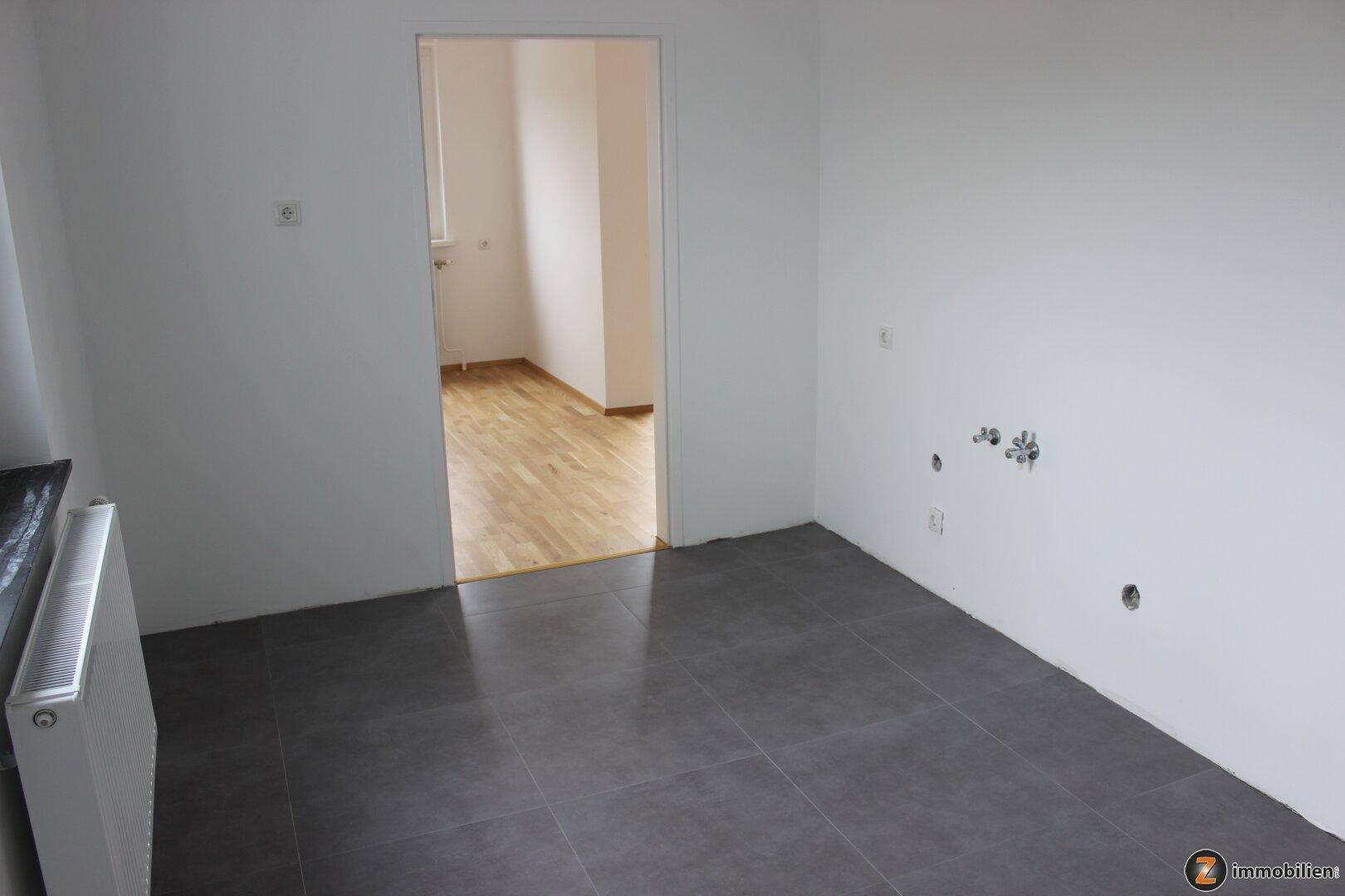 Leerer Küchenraum mit grauen Fliesen und vorbereiteten Anschlüssen, angrenzend an einen Raum mit Parkett.