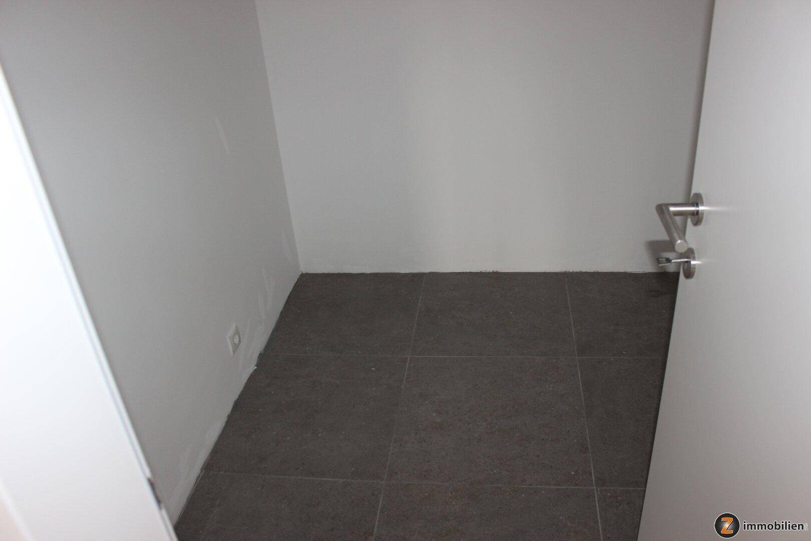 Leerer Abstellraum mit dunklem Fliesenboden und weißen Wänden, ideal für zusätzlichen Stauraum.