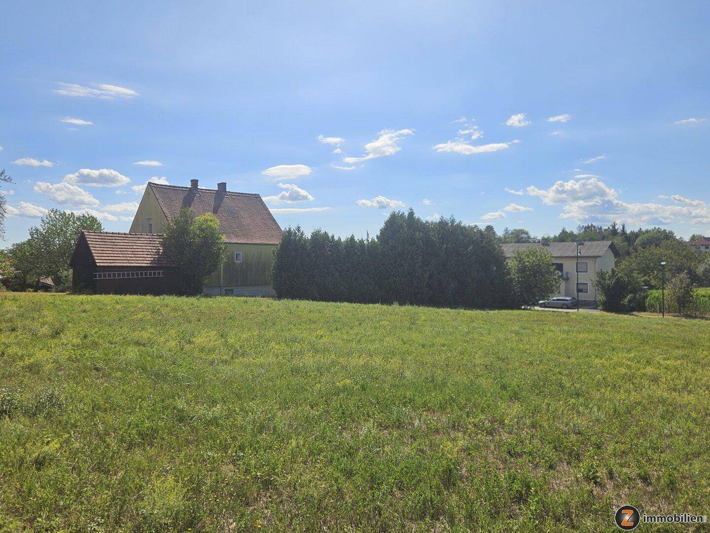 Grünes Grundstück mit Blick auf ein traditionelles Haus und dichte Bäume unter blauem Himmel.