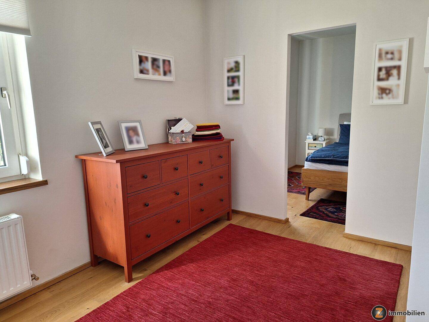 Schlafzimmer mit Kommode, rotem Teppich und Zugang zu einem weiteren Raum.