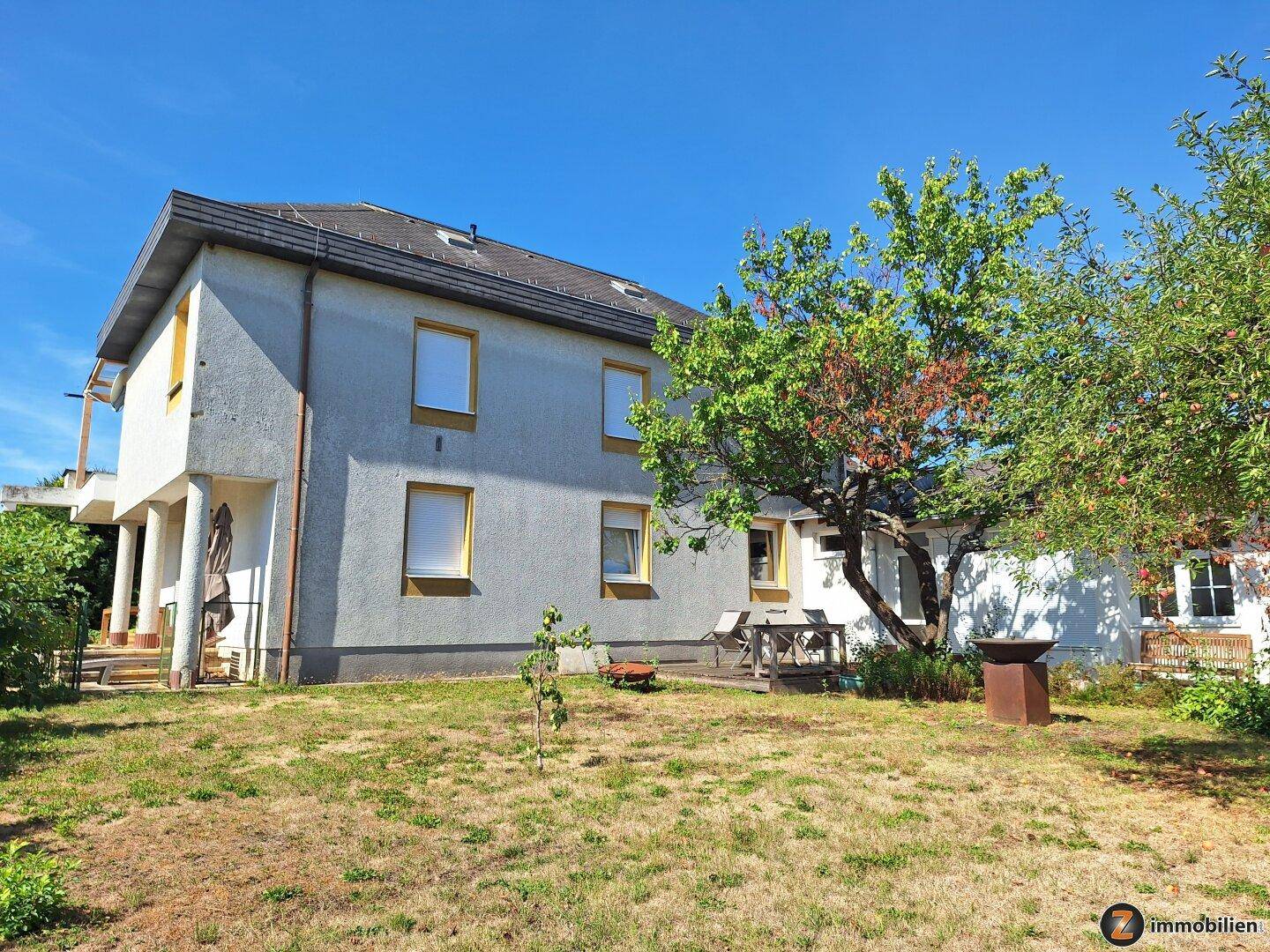 Rückansicht des Hauses mit Garten, Rasenfläche, Bäumen und einer kleinen Terrasse.