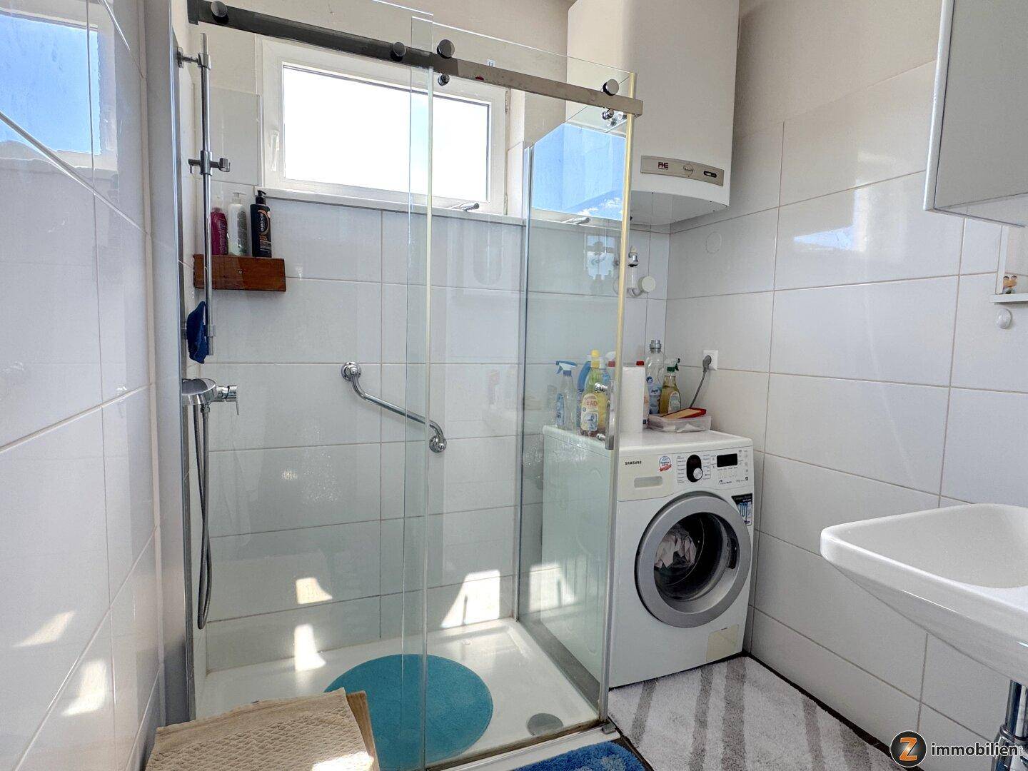 Helles Badezimmer mit Duschkabine, Waschmaschine und Fenster, das viel Tageslicht hereinlässt.