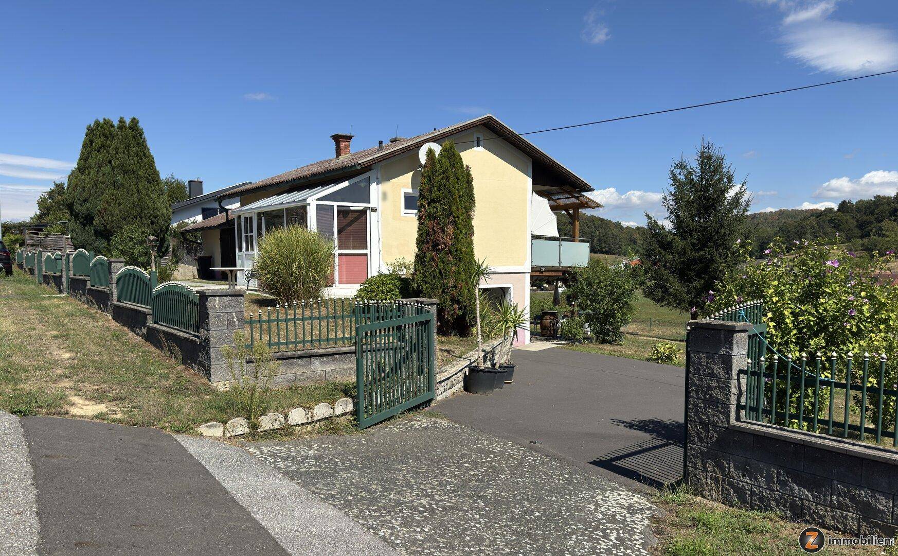 Einfamilienhaus mit gelber Fassade, Wintergarten, Balkon und gepflegtem Gartenbereich unter blauem Himmel.