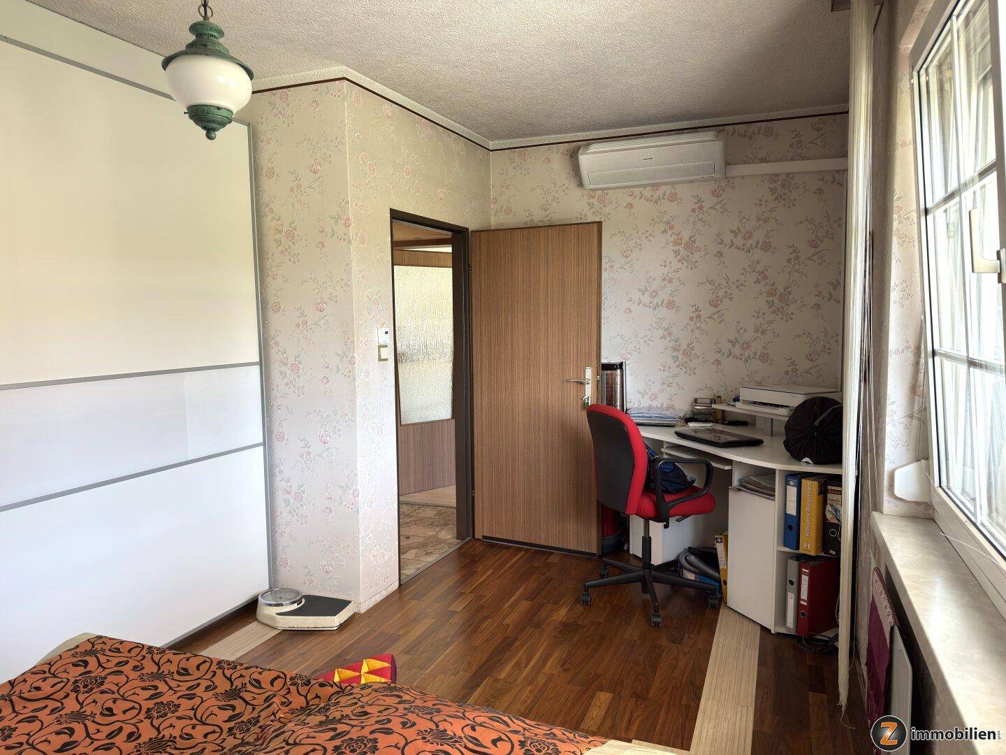 Schlafzimmer mit Schreibtisch, Schrank und Zugang zu einem weiteren Raum, ausgestattet mit Klimaanlage.