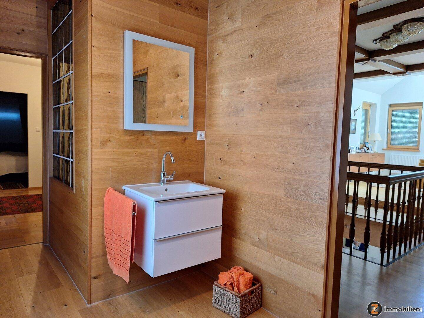 Modernes Badezimmer mit Einzelwaschbecken, Holzverkleidung und beleuchtetem Spiegel.