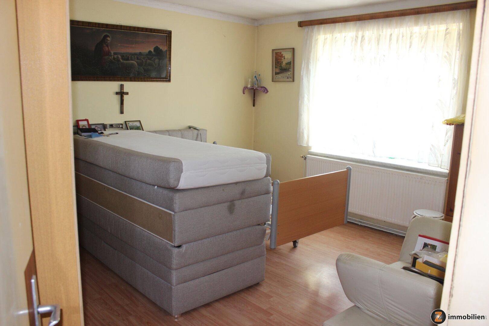Schlafzimmer mit gestapelten Matratzen, Heizkörper unter dem Fenster und religiöser Dekoration.