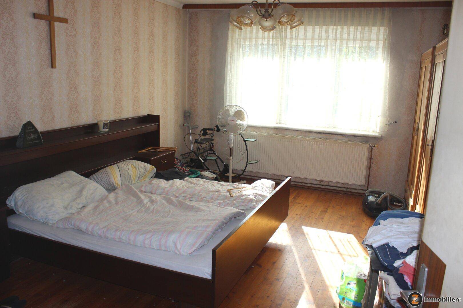 Gemütliches Schlafzimmer mit Holzbett, Ventilator und viel Sonnenlicht auf dem Holzboden.