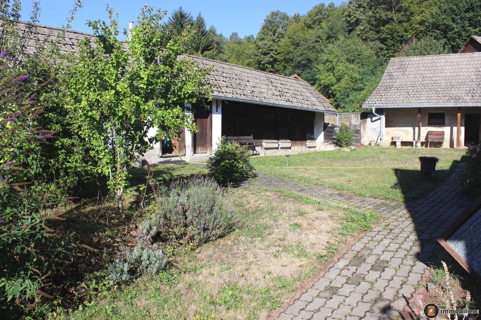 Garten mit gepflastertem Weg, Bäumen und zwei Nebengebäuden in ländlicher Umgebung.
