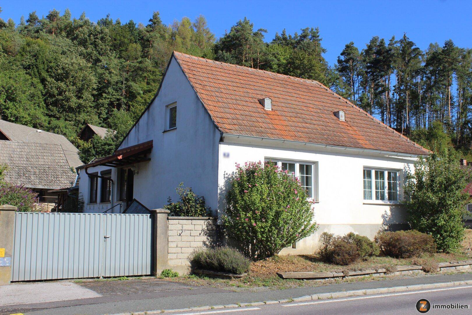 Weißes Einfamilienhaus mit rotem Ziegeldach, umgeben von Bäumen und einem Holzzaun.