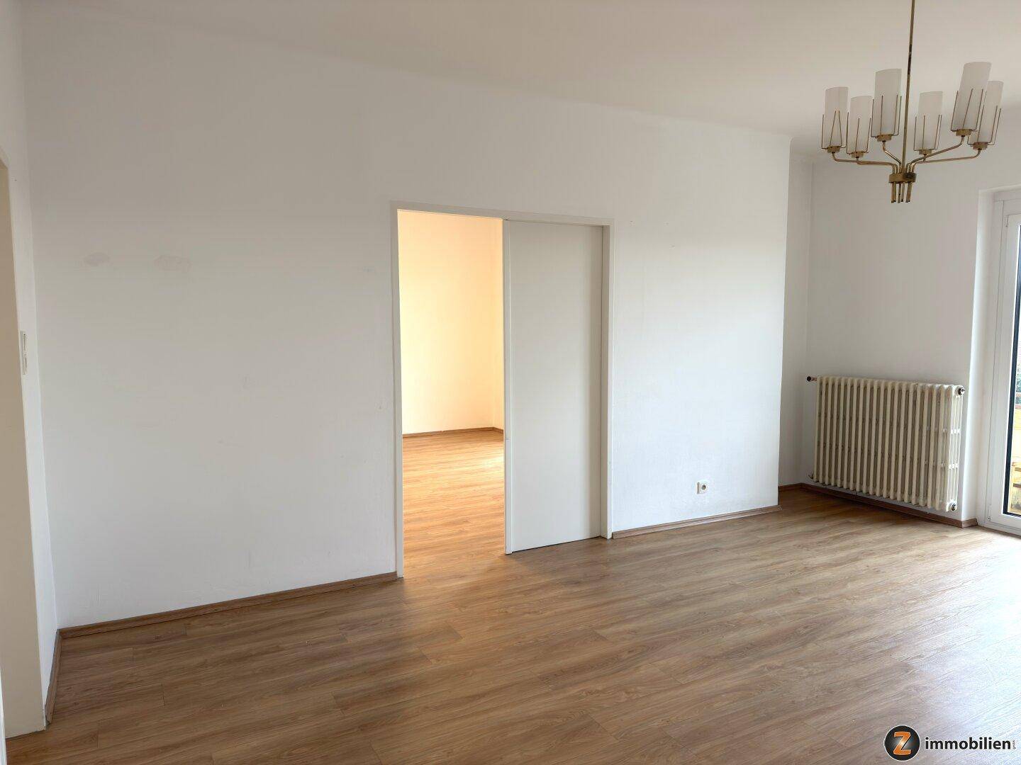 Helles Wohnzimmer mit Laminatboden und einer Schiebetür, die zu einem weiteren Raum führt.