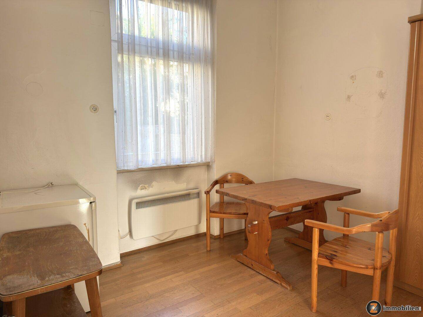 Einfaches Zimmer mit Holzboden, einem Tisch und Stühlen, sowie einem Fenster mit Vorhängen.
