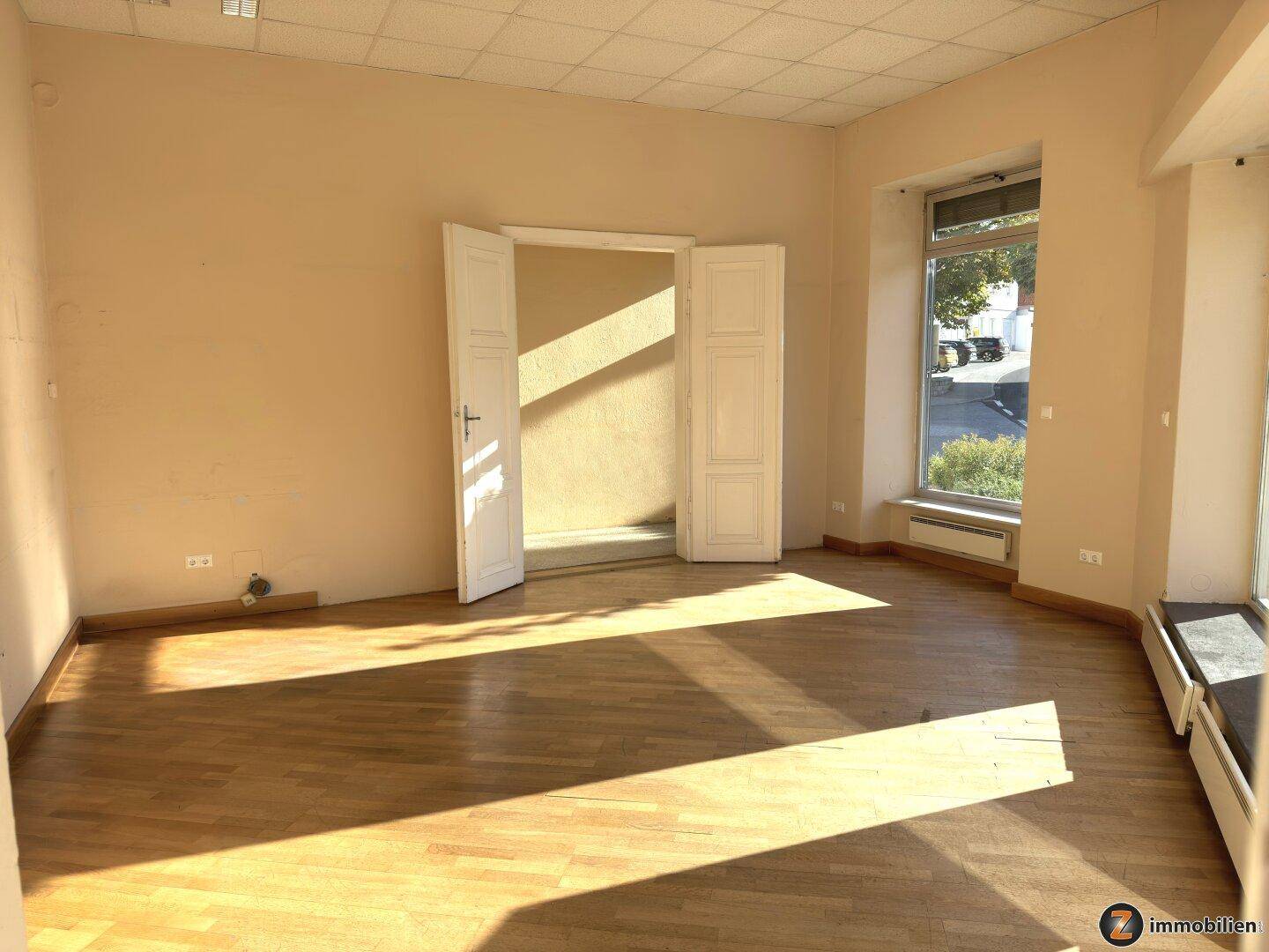 Geräumiger Raum mit Parkettboden und einem großen Fenster, das viel Sonnenlicht hereinlässt.
