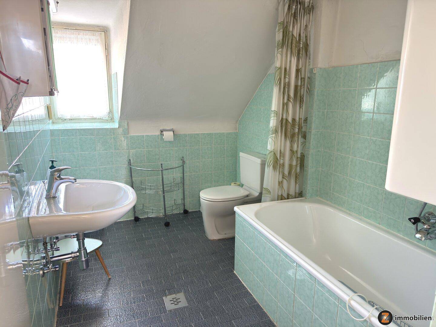 Badezimmer mit mintgrünen Fliesen, Badewanne, Waschbecken und Toilette unter einer Dachschräge.