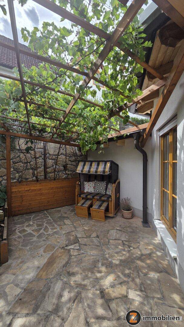 Überdachte Terrasse mit Steinboden, Sitzgelegenheit und Weinreben-Pergola.