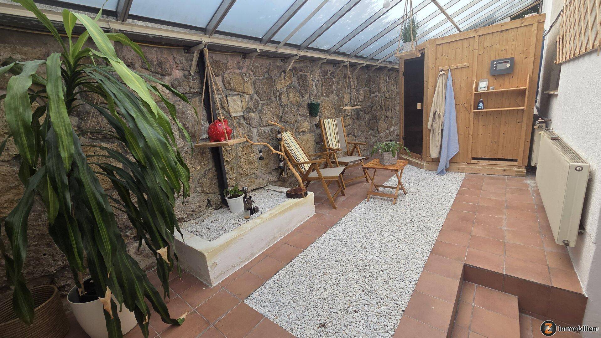 Entspannungsbereich im Wintergarten mit Sauna, Steinwand und Kiesboden.