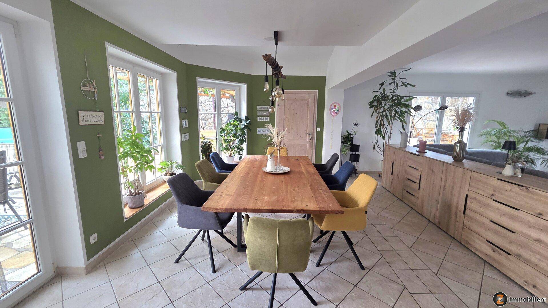 Heller Essbereich mit großem Holztisch, farbigen Stühlen und Zugang zur Terrasse.