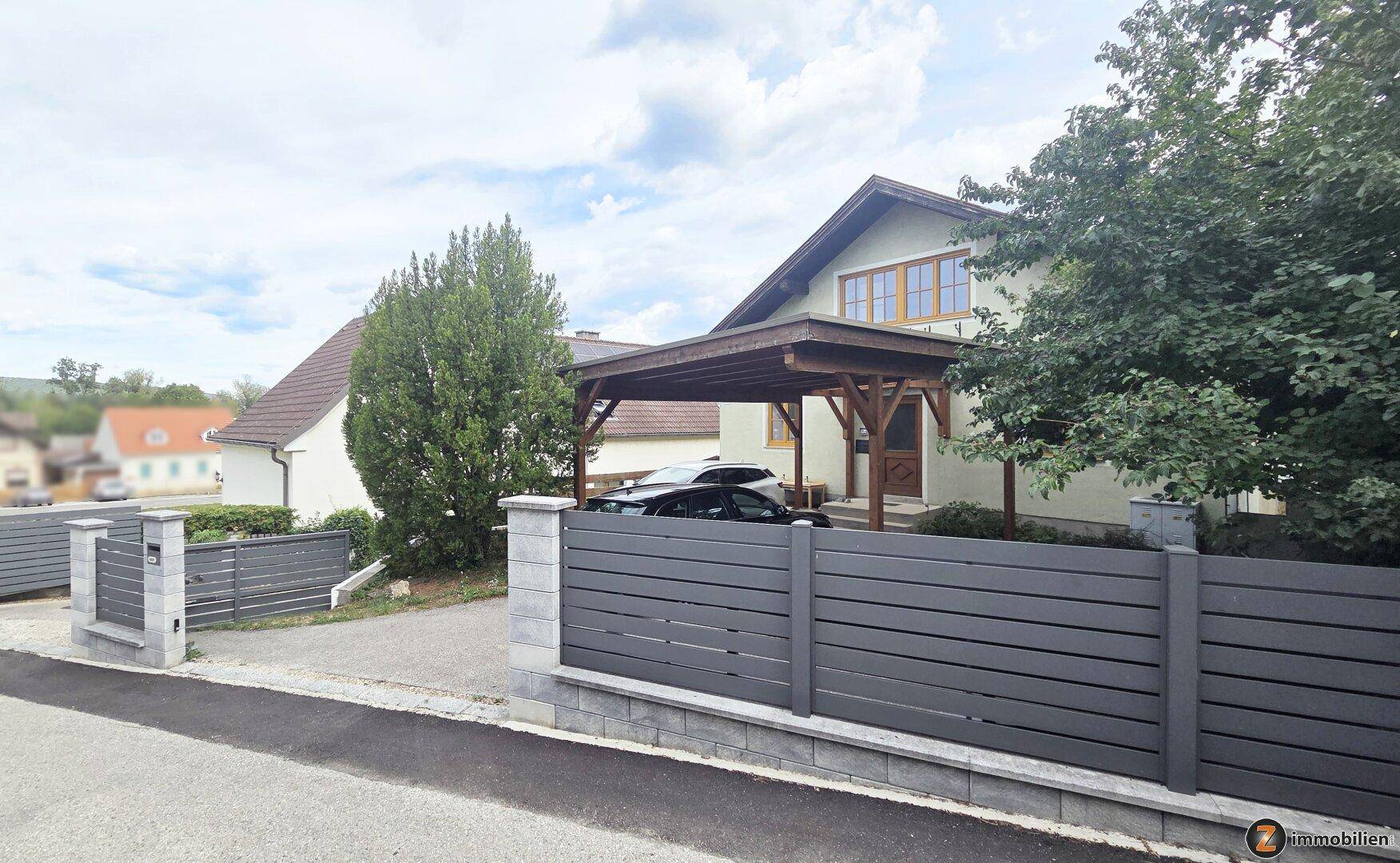 Einfamilienhaus mit modernem Zaun, Carport und gepflegtem Vorgarten in ländlicher Umgebung.