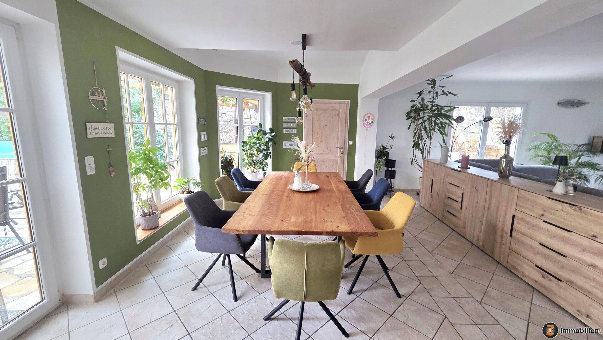 Heller Essbereich mit Holztisch, farbigen Stühlen und Zugang zur Terrasse, umgeben von Pflanzen.