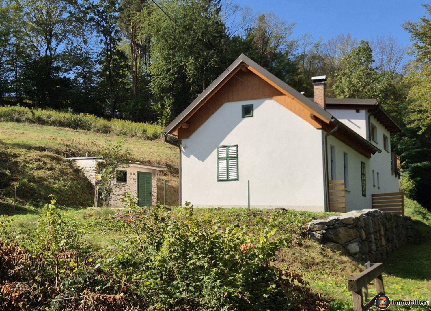 Weißes Landhaus mit grünen Fensterläden und Holzelementen, eingebettet in eine grüne Hügellandschaft.