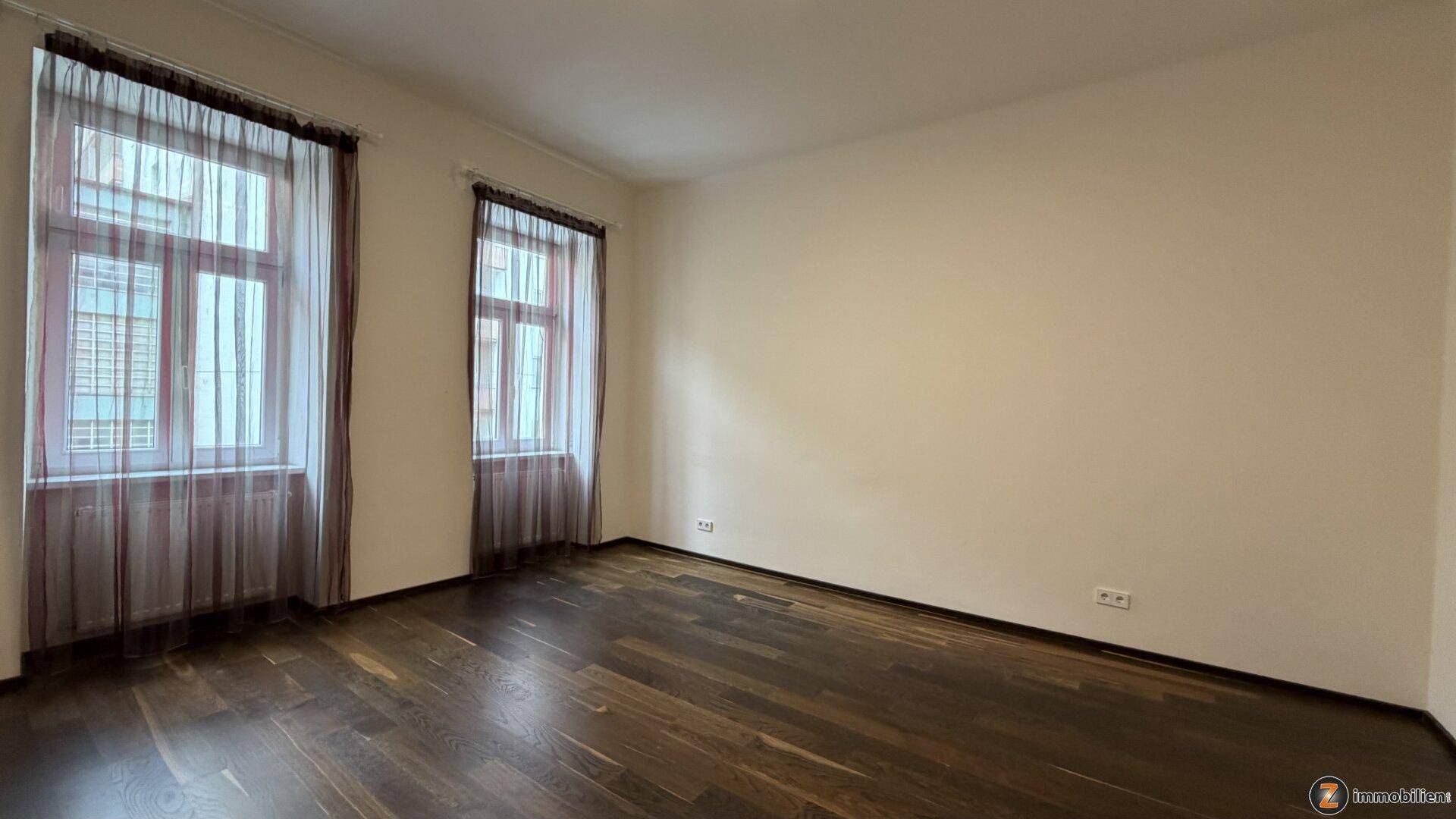Geräumiger, heller Raum mit dunklem Holzboden und zwei großen Fenstern.